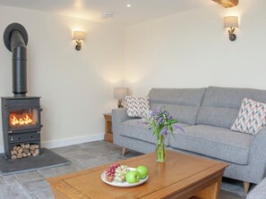 A living room with a wood-burning stove, grey sofa, wooden coffee table with fruit, and wall-mounted lamps.