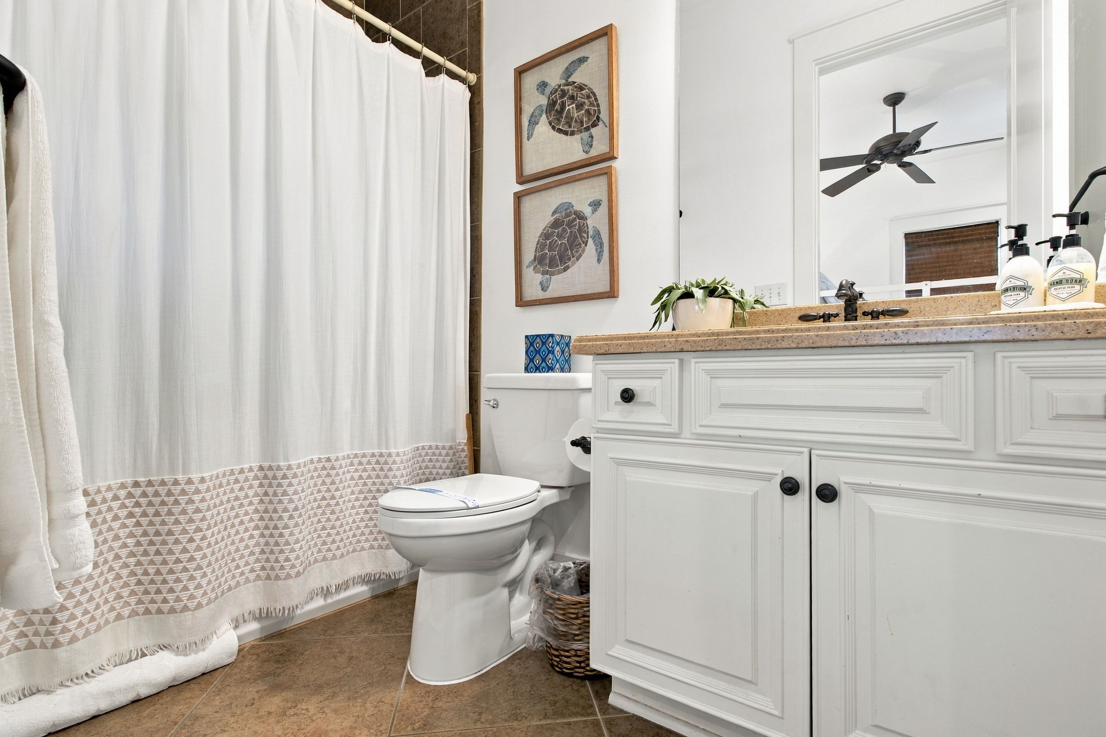 A bathroom with a toilet, a shower curtain, a sink with soap dispensers, and a ceiling fan.