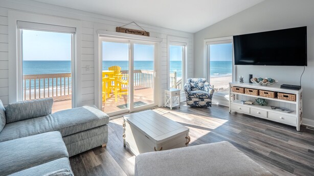 A spacious living room with a large TV, a sofa, a coffee table, and a view of the beach through large windows. - Keeping It Salty #101 Oceanfront Duplex, Private Beach Access, Dogs Allowed! (Surf City)