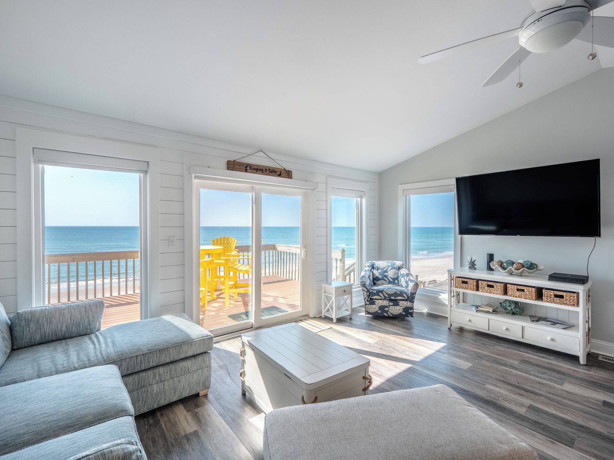 A spacious living room with a large TV, a sofa, a coffee table, and a view of the beach through large windows.