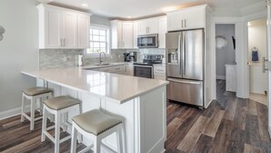 A modern kitchen with white cabinetry, a large island with stools, stainless steel appliances, and a herringbone wood floor. - Keeping It Salty #101 Oceanfront Duplex, Private Beach Access, Dogs Allowed! (Surf City)