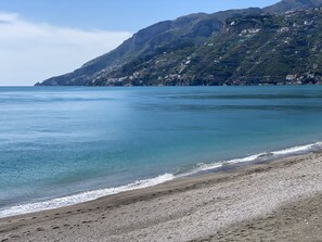 A coastal landscape with a pebble beach, clear blue water, and a hillside with buildings.