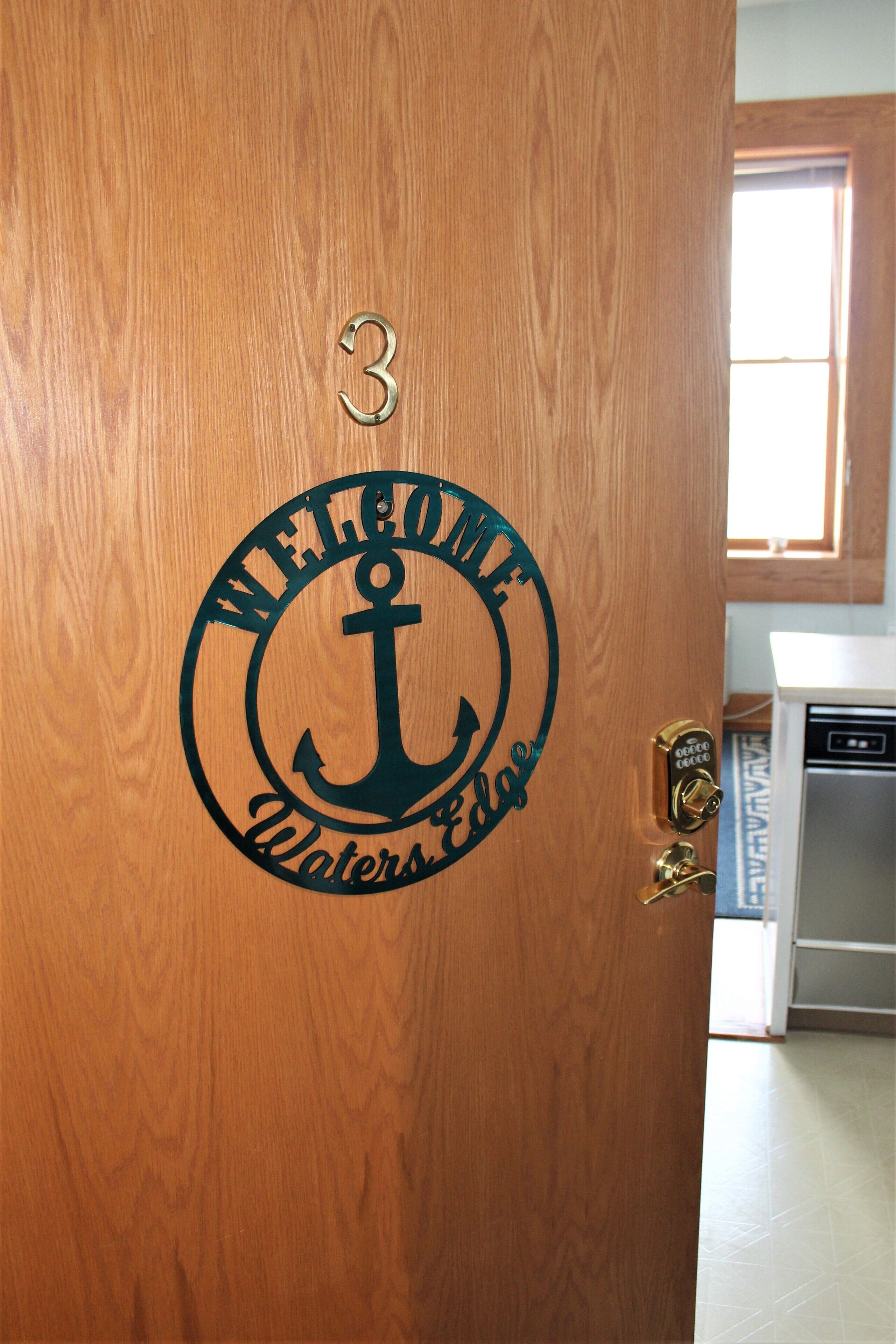 Una porta di legno con una targa di benvenuto che presenta un'ancora e il testo "Welcome Waters Edge".