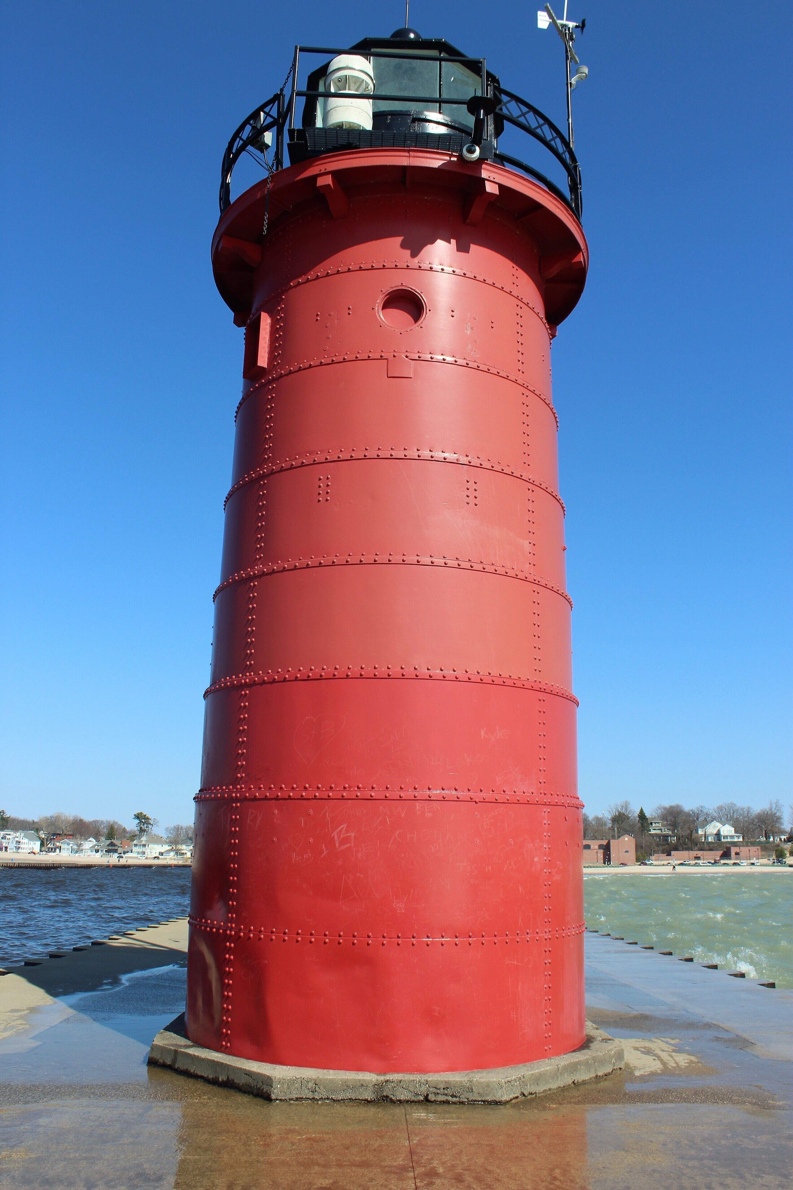 A red cylindrical lighthouse with a weather vane on top, situated by a waterfront with a clear blue sky.