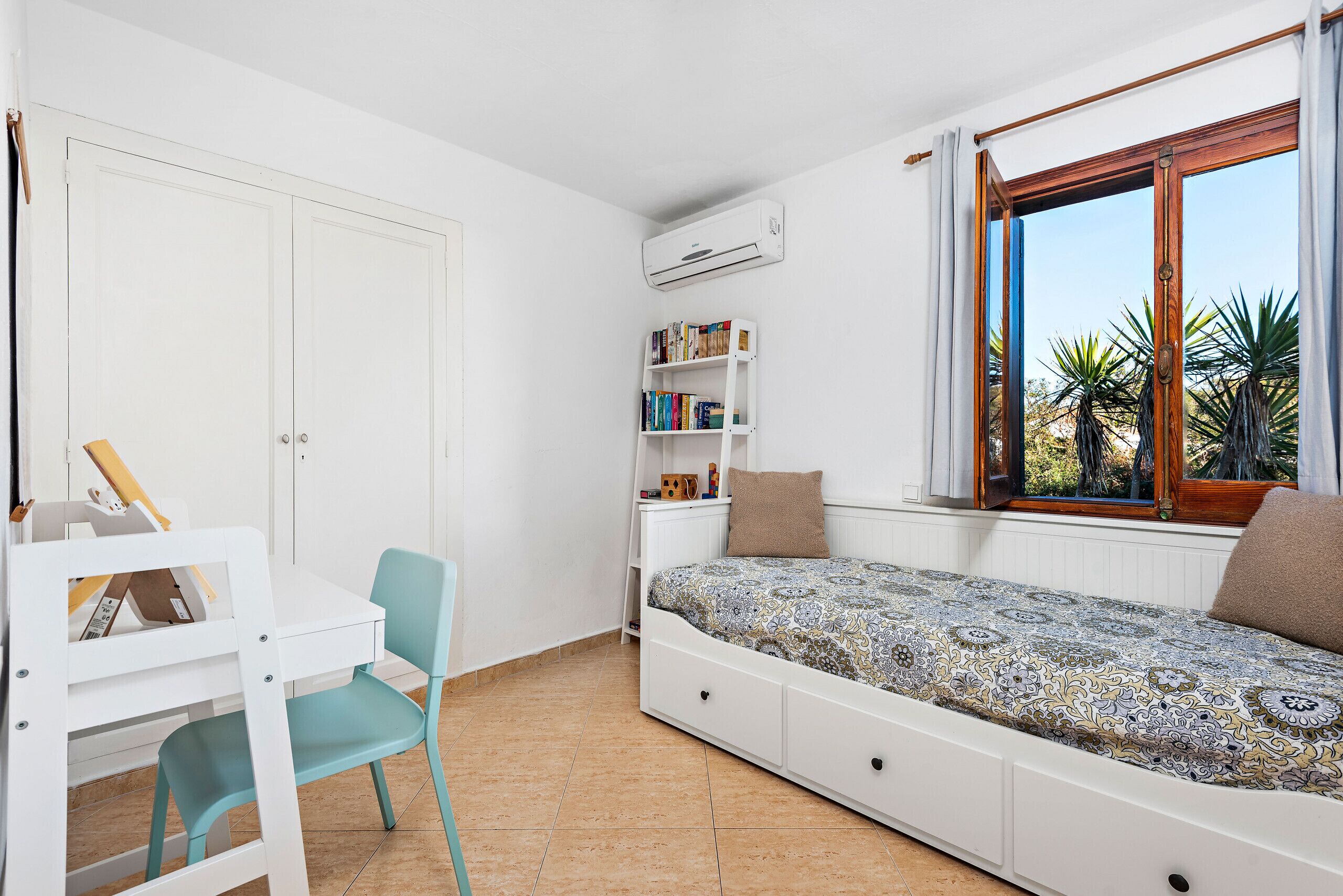 Una habitación pequeña con una cama, un escritorio, una silla, una estantería y un ventanal con cortinas.