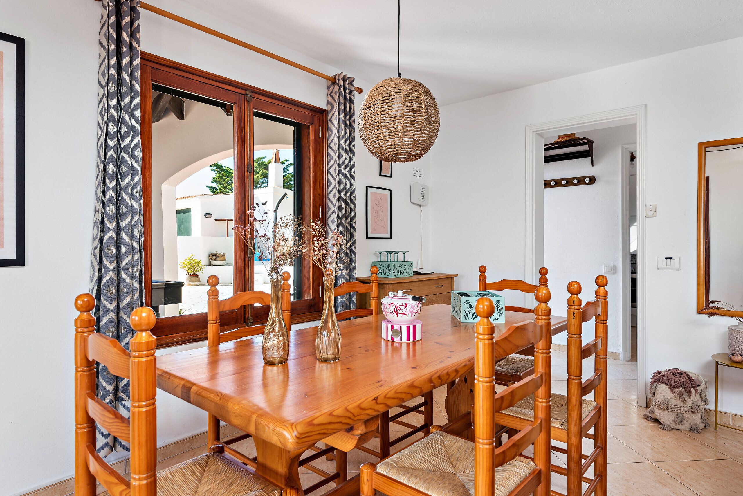A dining area with a wooden table and chairs, a hanging woven light fixture, and a view through an open doorway to an outdoor space.