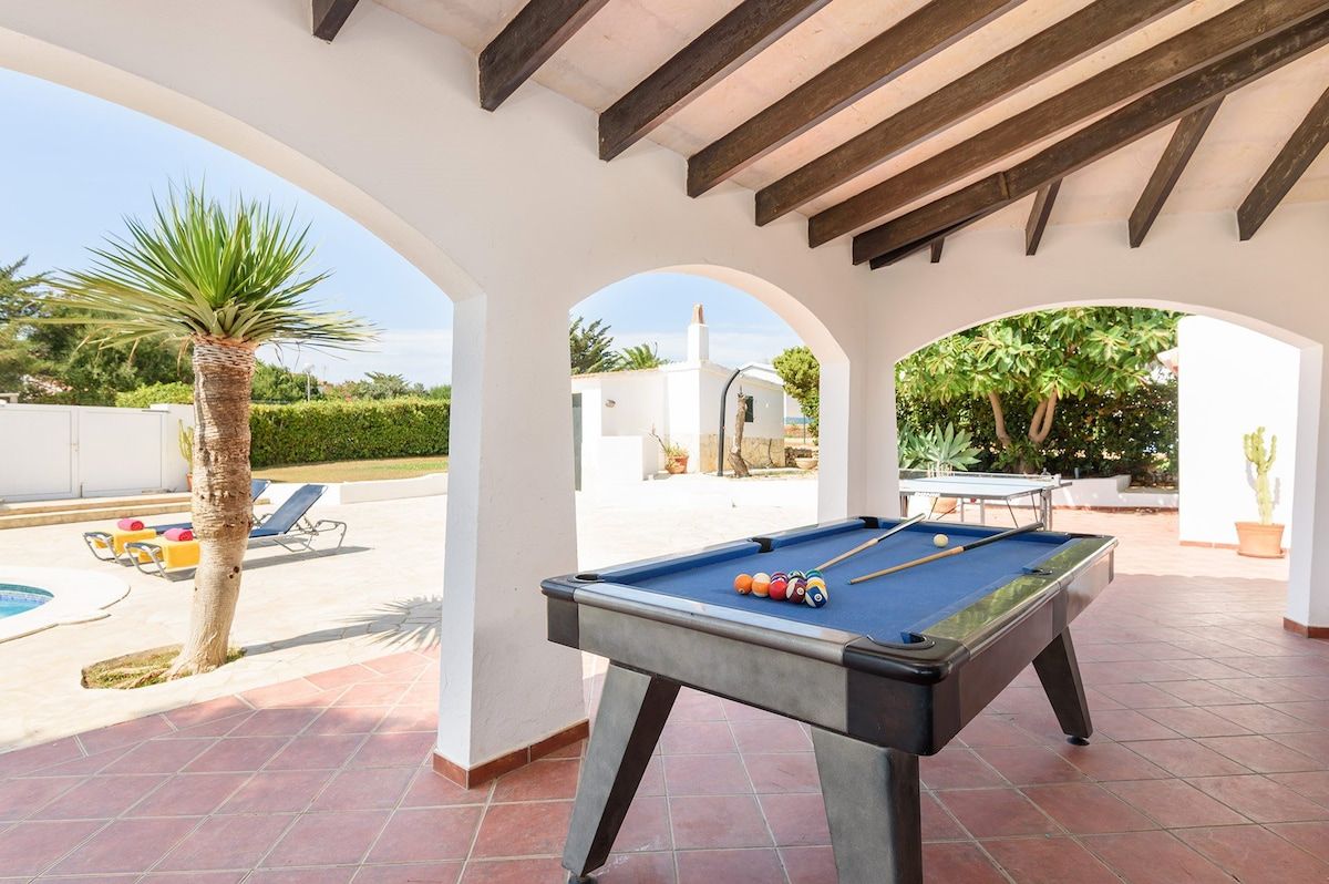 Zona de piscina exterior con mesa de billar, sillas de descanso y una palmera.
