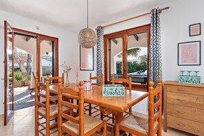 Un comedor con mesa y sillas de madera, una lámpara colgante y vistas amplias a través de ventanas grandes.