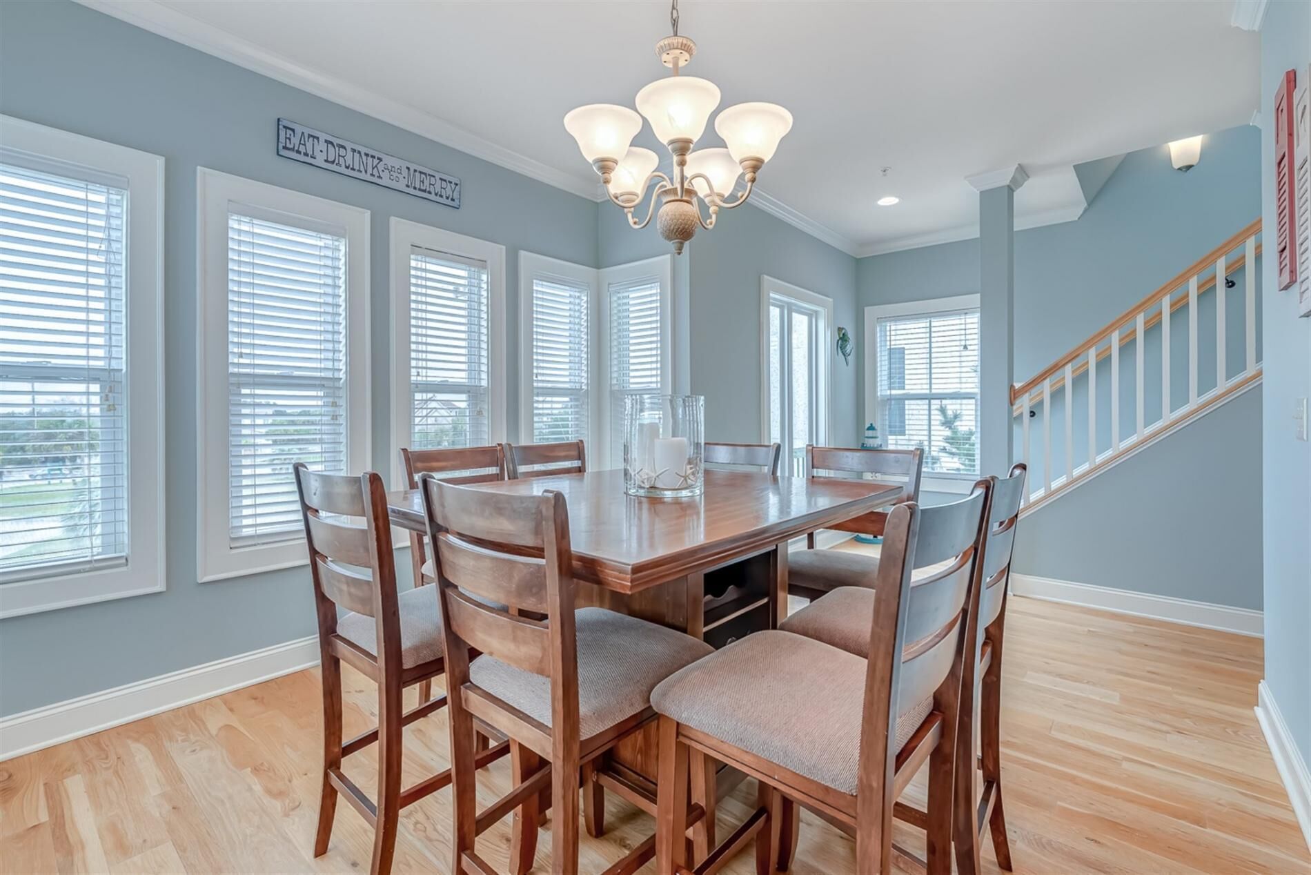A dining area with a wooden table and chairs, a chandelier, and large windows with blinds.