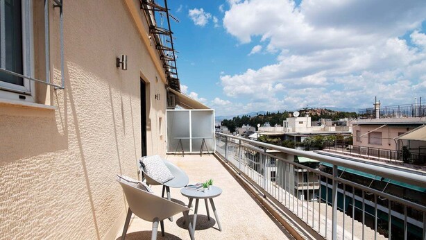 Un balcon avec deux chaises et une petite table, d’où l’on peut admirer un paysage urbain avec ses bâtiments et un ciel dégagé.