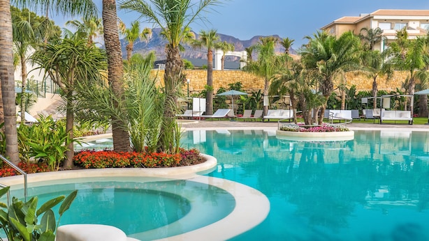 Zona de piscina con sillones de descanso, palmeras y montañas al fondo.