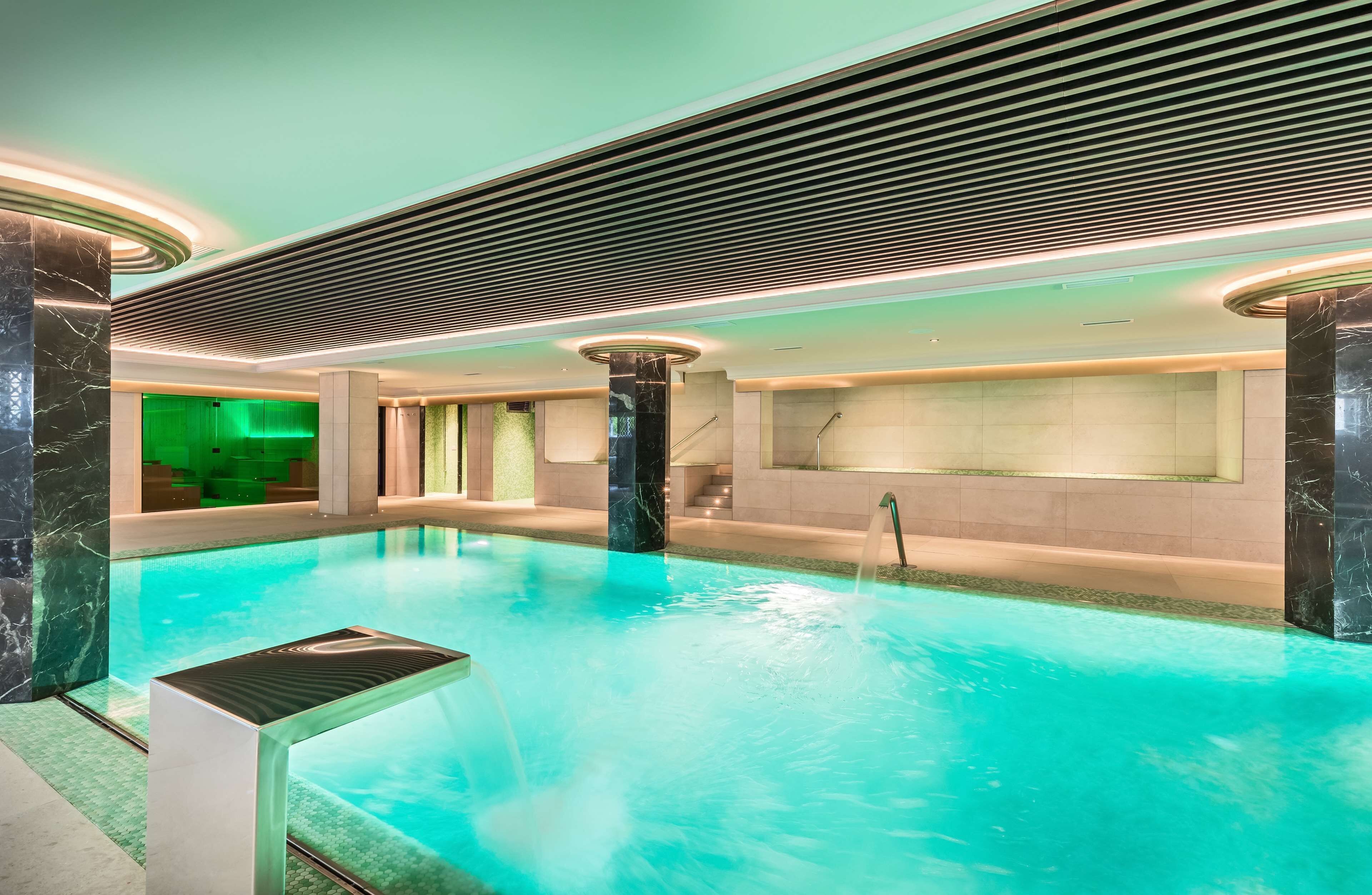 A modern indoor swimming pool with a central column, tiled walls, and a ceiling with a linear light fixture.