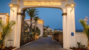 Entrada a Barceló La Nucía Palms con un camino despejado, palmeras y un cartel que indica que es un hotel.
