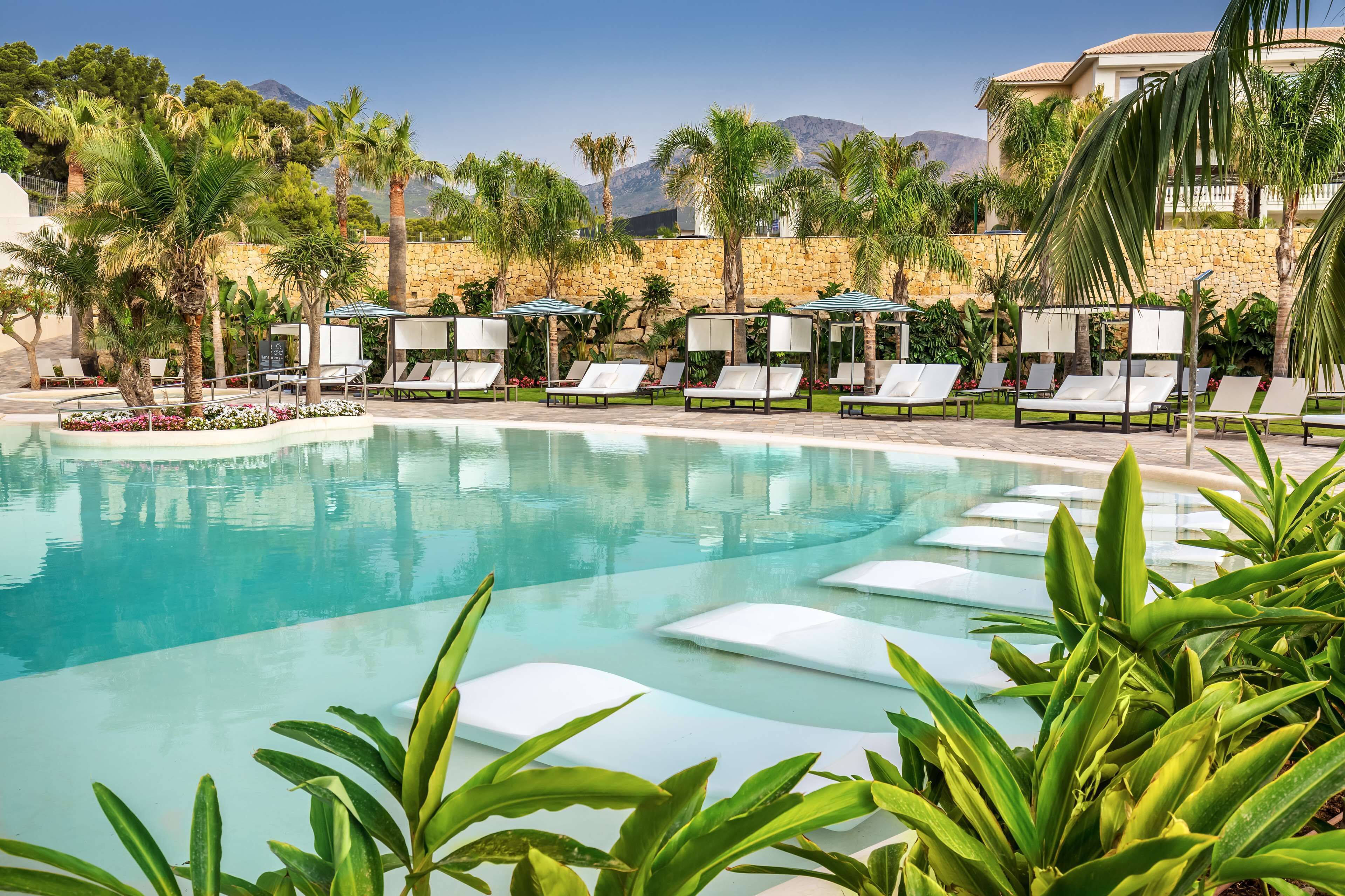 A pool area with lounge chairs and palm trees, surrounded by a stone wall and a mountain in the background.