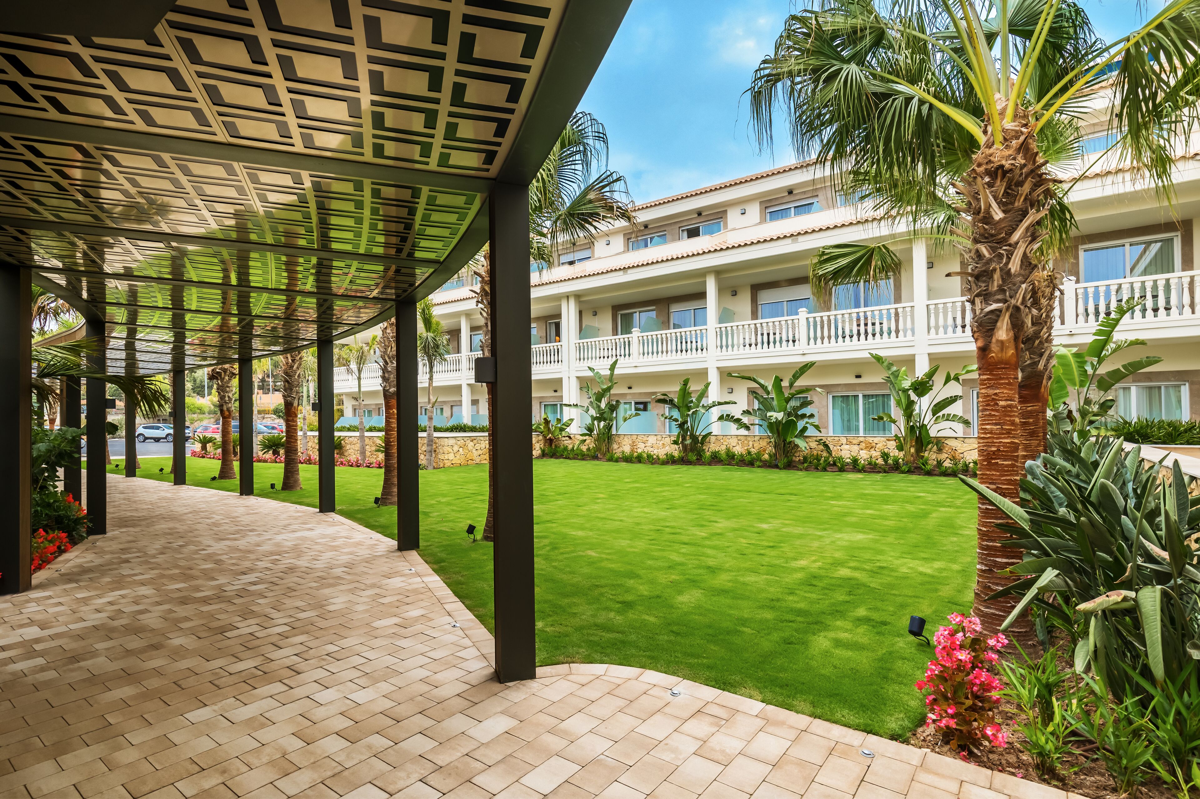 A covered walkway with a tiled floor, a well-maintained lawn, and a multi-story building with balconies in the background.