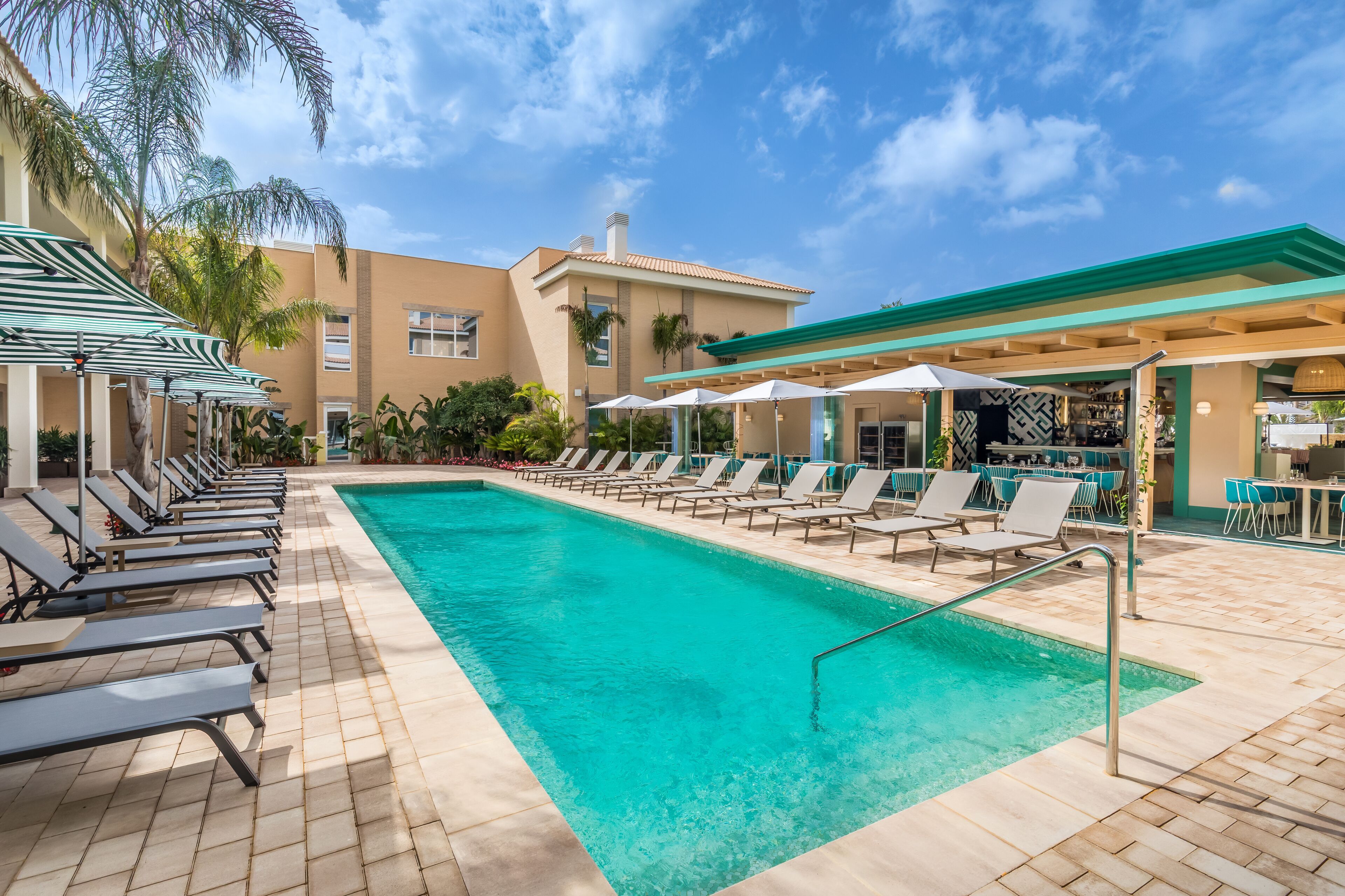 A pool area with lounge chairs and umbrellas, a bar, and a building with a green roof.