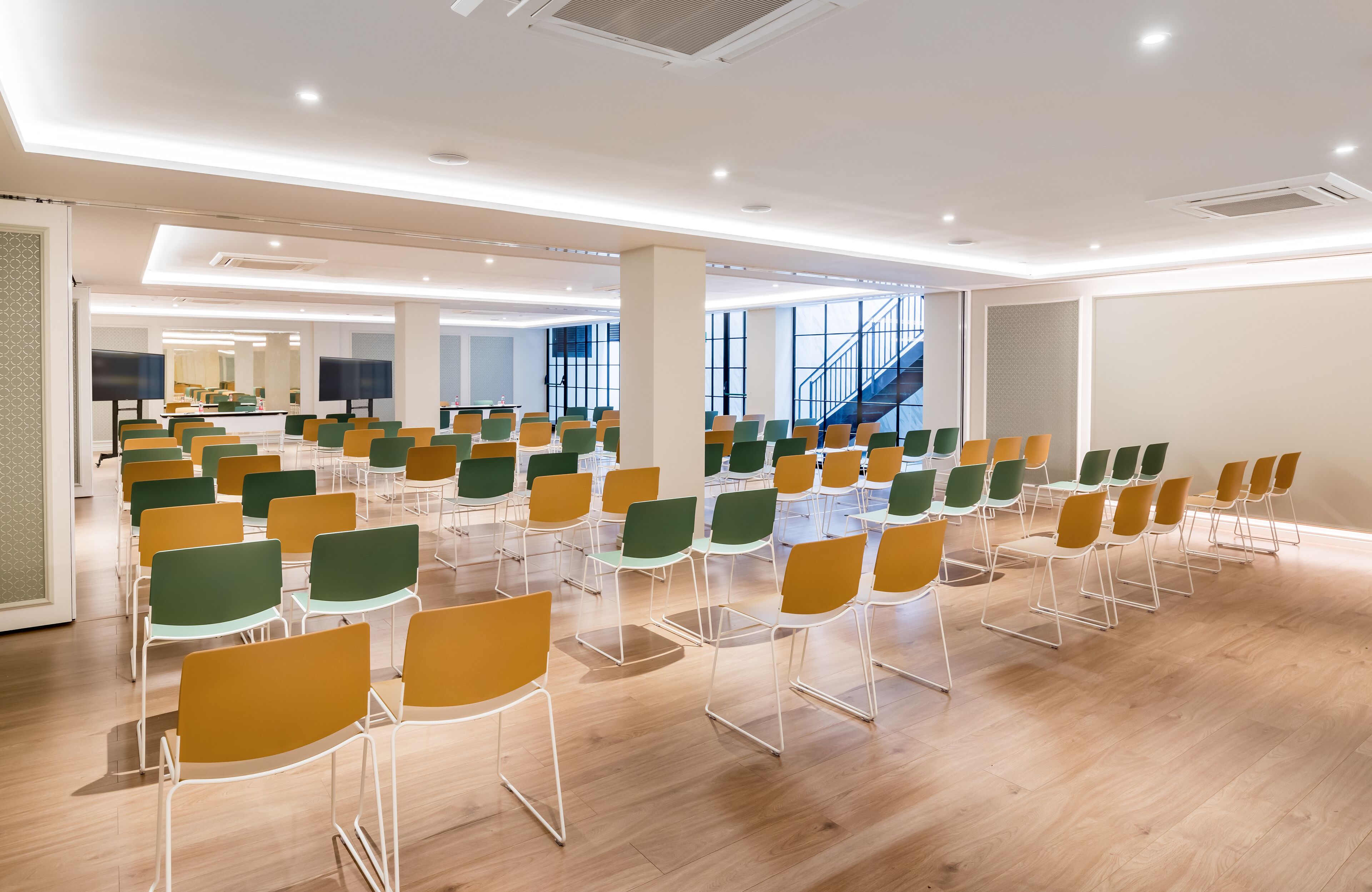 A spacious conference room with rows of chairs, large windows, and a staircase in the background.