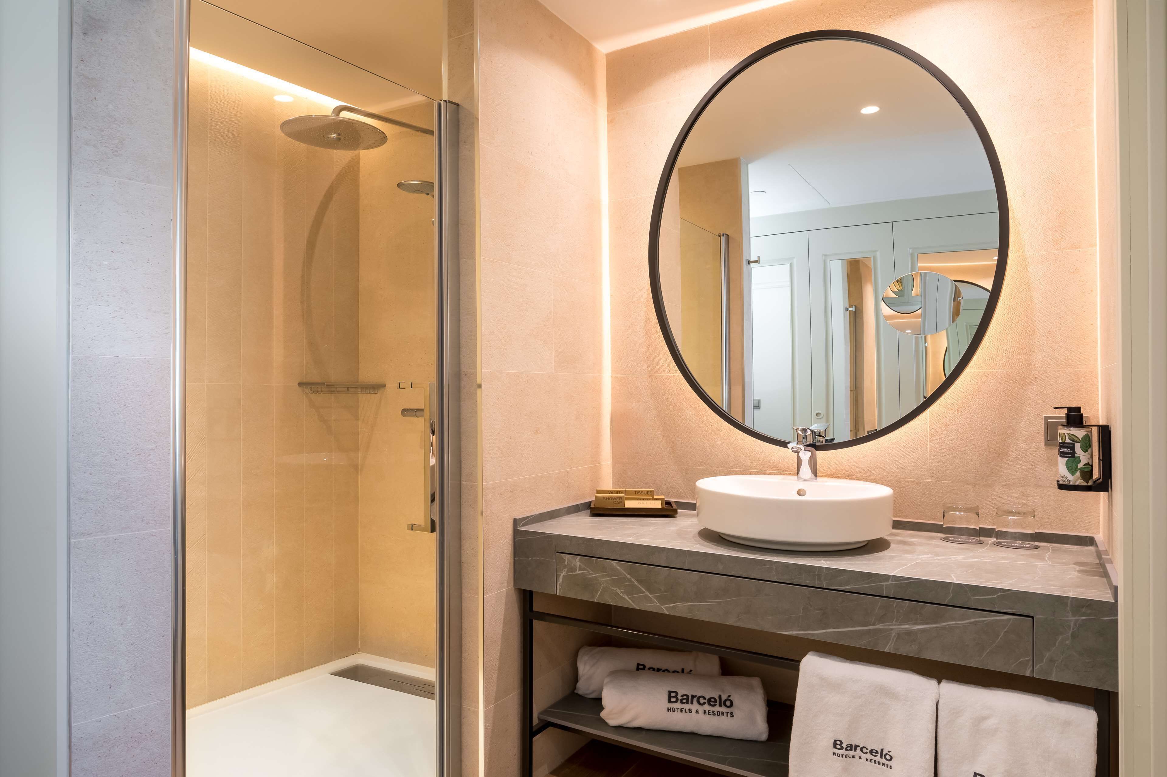 A modern bathroom with a glass-enclosed shower, a round mirror, a white sink, and a marble countertop. There are towels with the hotel name on the shelf.