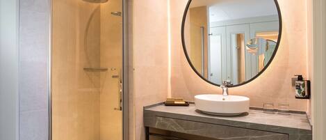 A modern bathroom with a glass-enclosed shower, a round mirror, a white sink, and a marble countertop. There are towels with the hotel name on the shelf.