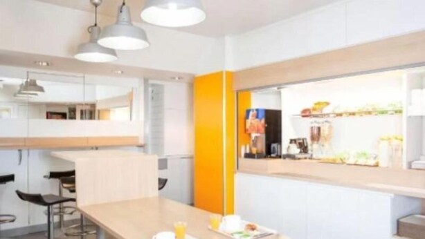 A modern kitchen with a breakfast bar, bar stools, and a bright orange accent wall. - Premiere Classe Arras - Tilloy Les Mofflaines (Tilloy-Les-Mofflaines)