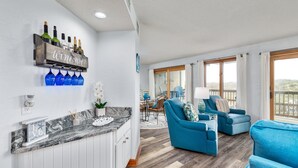 Uma sala de estar moderna com mobiliário azul, uma área de cozinha com bancada de mármore e um espaço bar com garrafas de vinho.