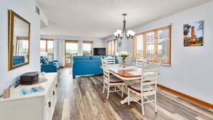 Uma sala de estar moderna com um sofá azul, uma área de jantar com uma mesa e cadeiras de madeira, e grandes janelas com persianas.