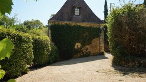 A stone building with a steeply pitched roof, surrounded by dense greenery and a gravel path leading to the entrance.