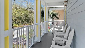 Une véranda couverte, avec des chaises blanches et une balustrade, donnant sur un jardin arboré et sur un bâtiment visible au loin.