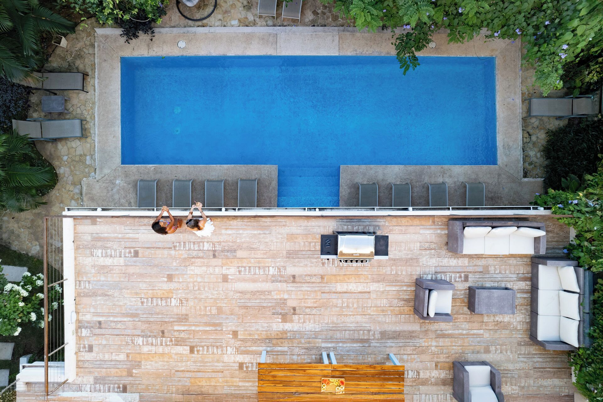 An aerial view of a rectangular swimming pool with a wooden deck surrounding it, equipped with multiple lounge chairs and a barbecue area.