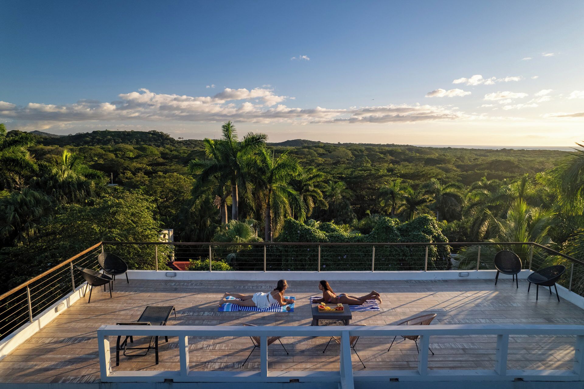 A rooftop terrace with two sun loungers, a table with a drink, and a view of a lush landscape with palm trees.