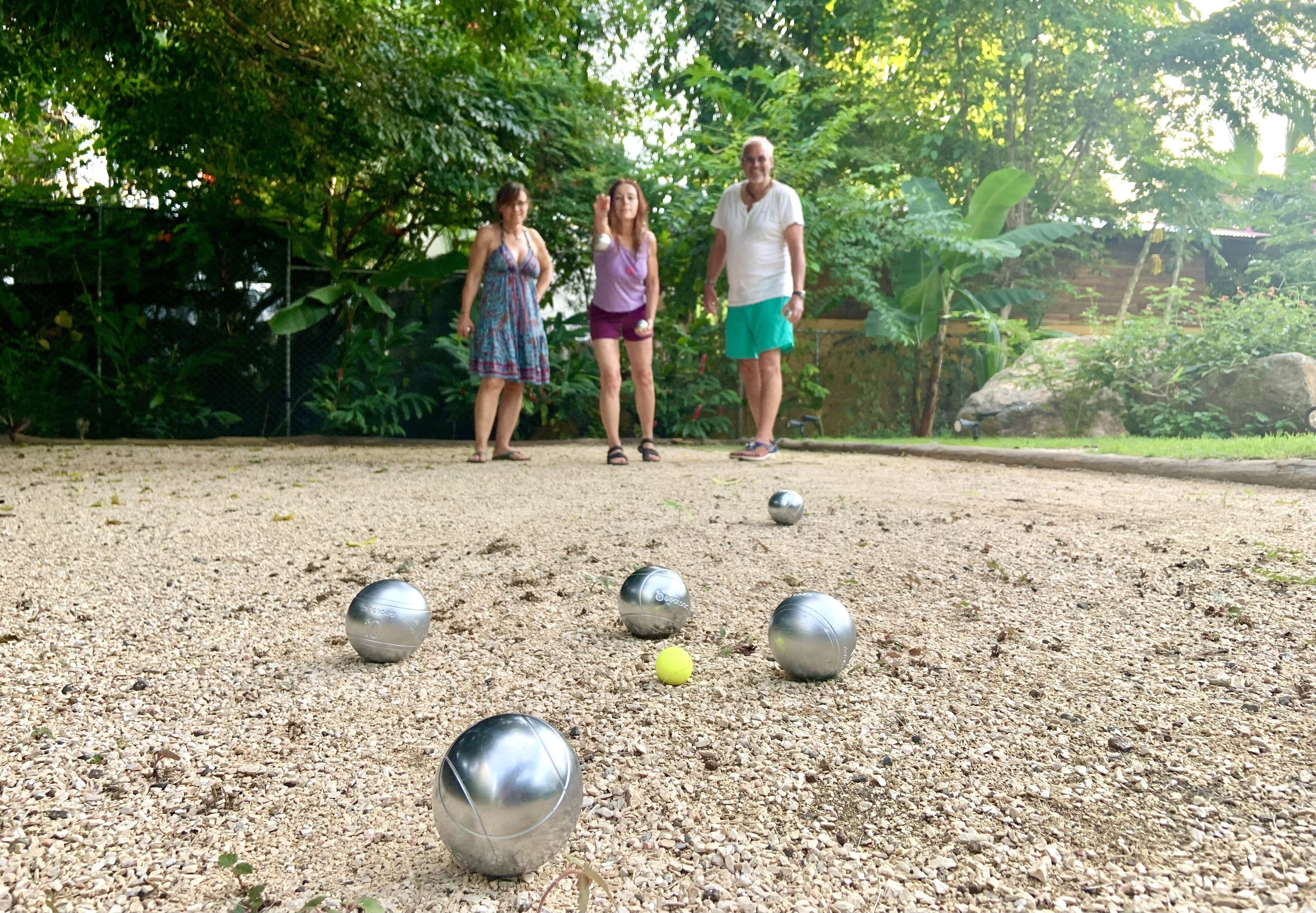 A group of people playing a game with metallic balls on a gravel surface, surrounded by greenery.