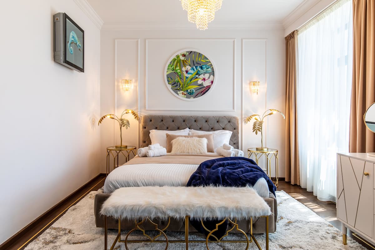 A bedroom with a tufted headboard, a round wall art piece, bedside lamps, a large mirror, and a view of the outdoors through sheer curtains.