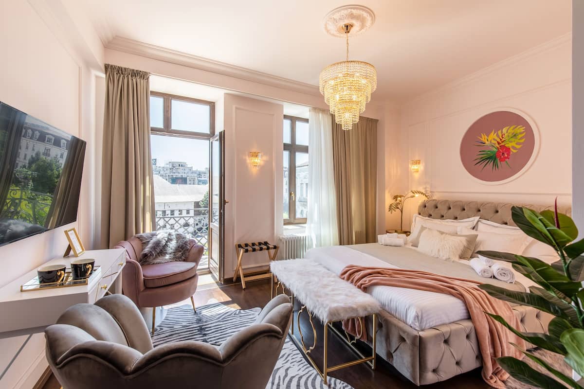 A hotel room with a bed, a desk, a chair, a bench, a TV, and a balcony.