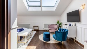 Un dormitori modern estil loft amb lluminàries laterals, un llit, una zona de seients amb un otomà blau, una cadira rosa i un televisor fixat a la paret.