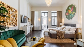 Habitació d'hotel moderna amb un sofà verd, una butaca groga, un llit amb una manta beix, una taula de cafè de vidre amb una espelma i un quadre gran de pássers a la paret.