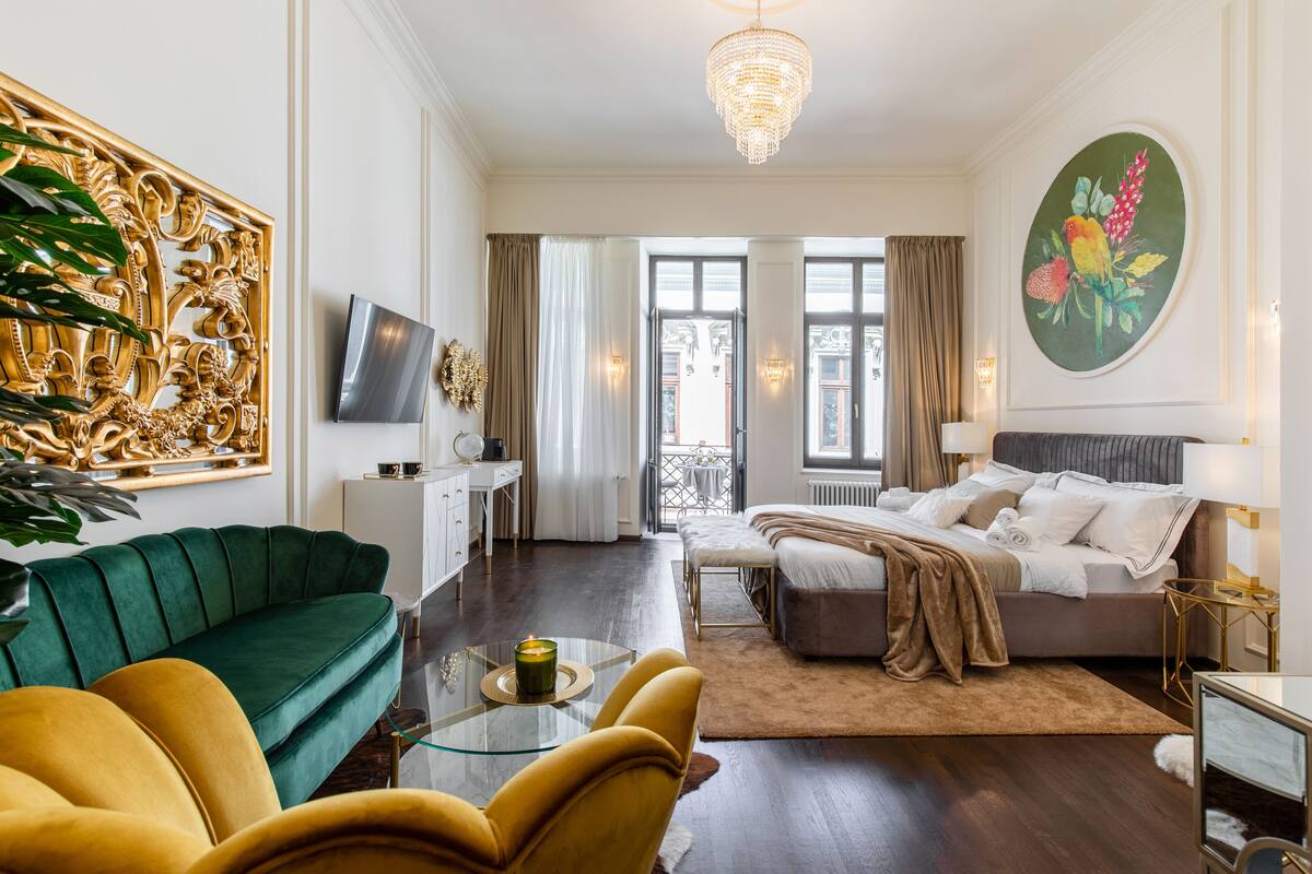 A modern hotel room with a green sofa, a yellow armchair, a bed with a beige blanket, a glass coffee table with a candle, and a large painting of birds on the wall.