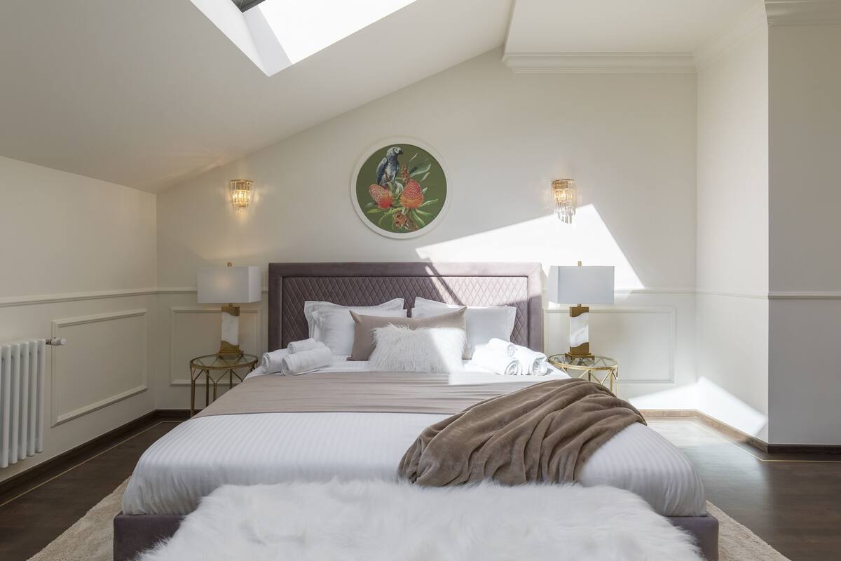 A bedroom with a large bed, two bedside lamps, a round painting, and a skylight.