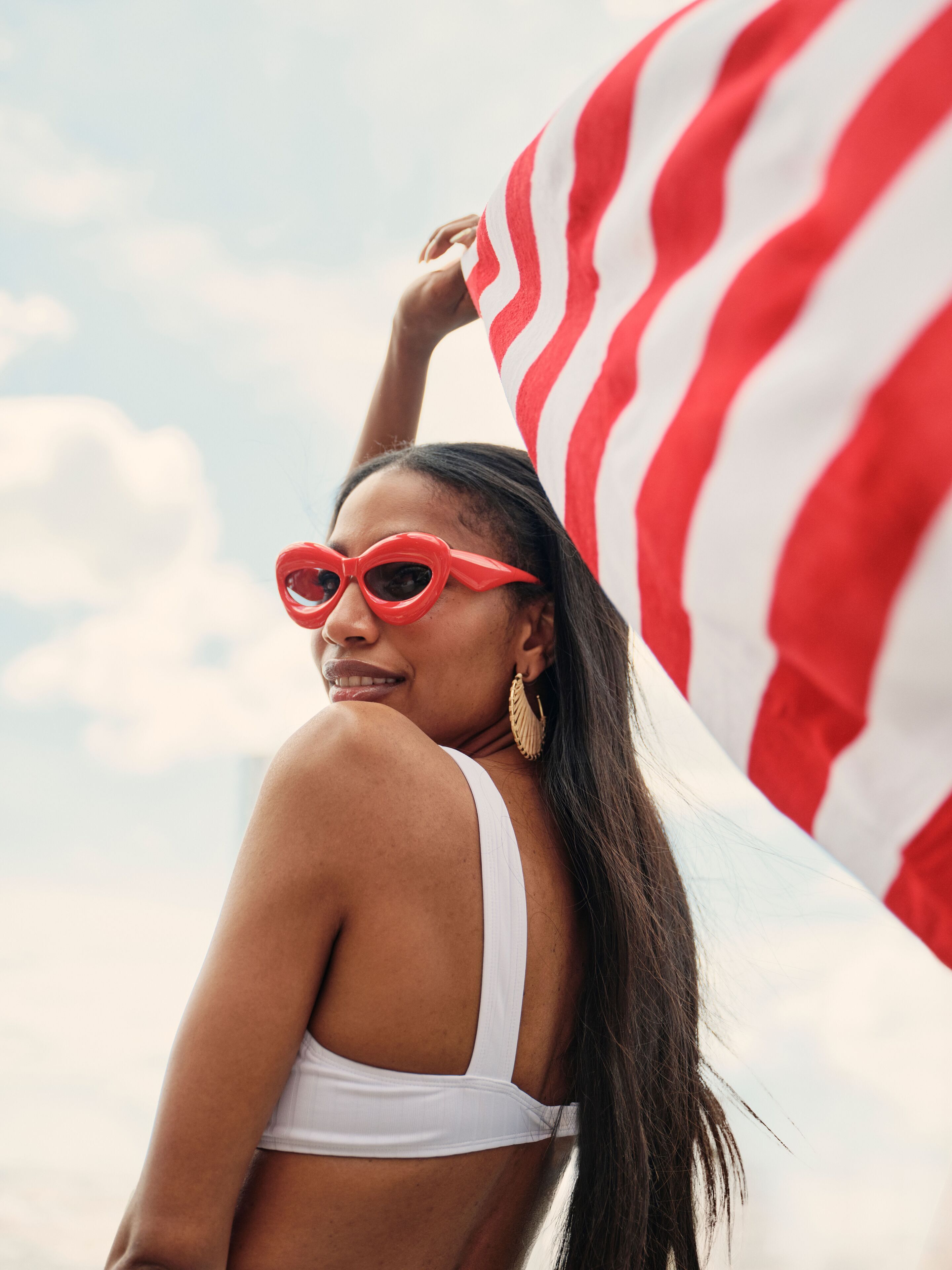 Una persona con un top bikini bianco e occhiali da sole rossi, con una grande bandiera americana sullo sfondo.