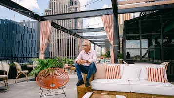 A man sitting on a white sofa with orange and white striped cushions, surrounded by wicker furniture and potted plants in an outdoor seating area with a city view.