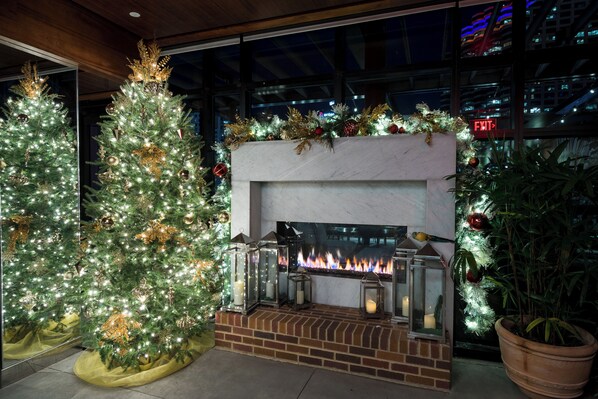 A fireplace with a brick surround, flanked by two Christmas trees adorned with lights and ornaments. - Virgin Hotels New Orleans (New Orleans)