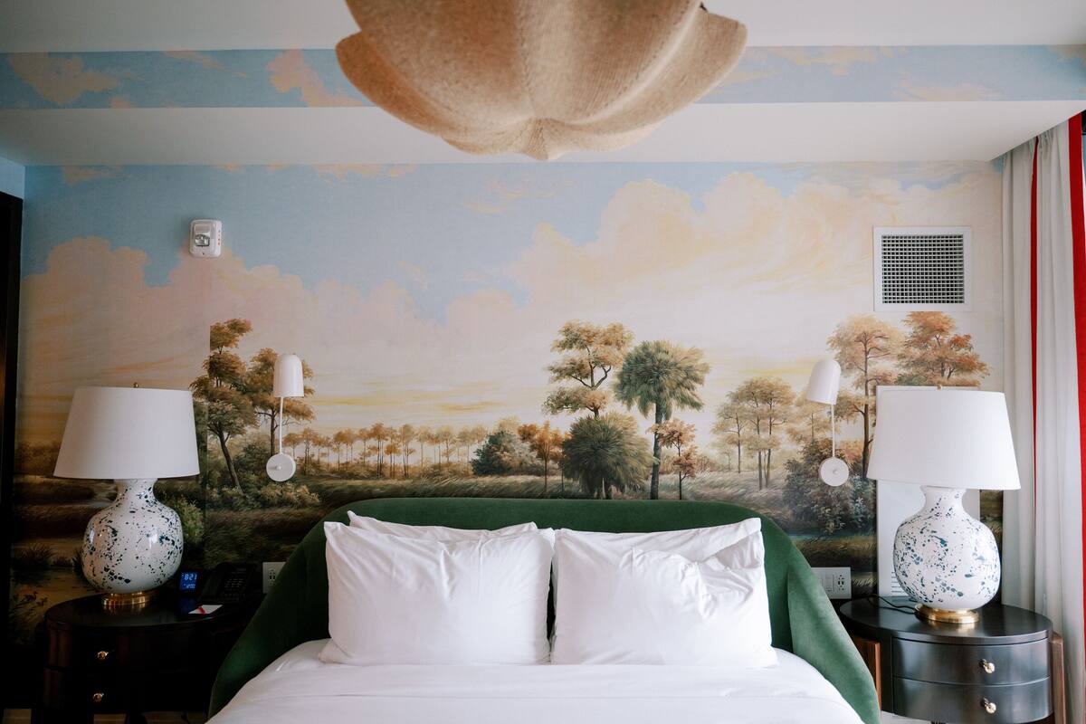 Une chambre d’hôtel avec un lit, deux tables de chevet, un plaid vert et une fresque représentant un paysage.
