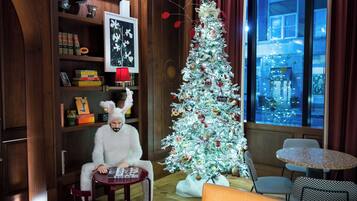 A cozy room with a Christmas tree, a bookshelf with books and decorative items, a window with a view of the city, and a small round table.