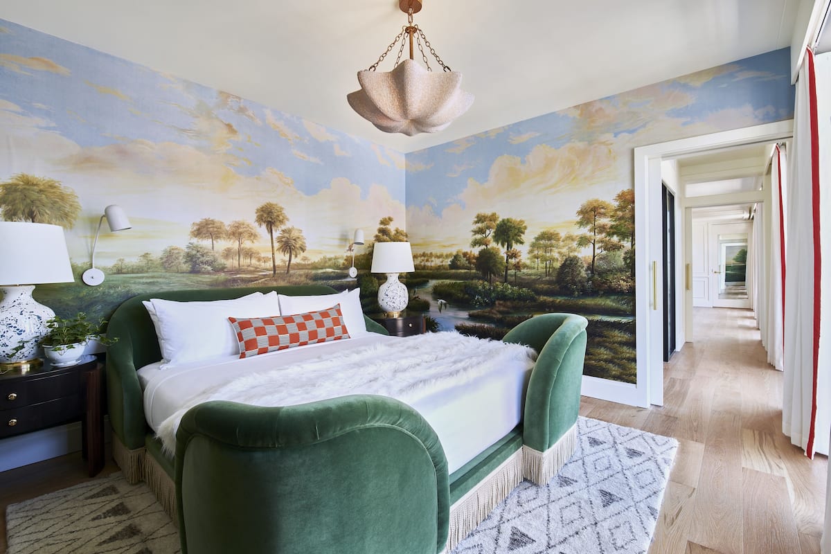 Une chambre à coucher ornée d’une grande fresque représentant un paysage, avec un lit recouvert d’un tissu vert, deux lampes de chevet et un luminaire suspendu.