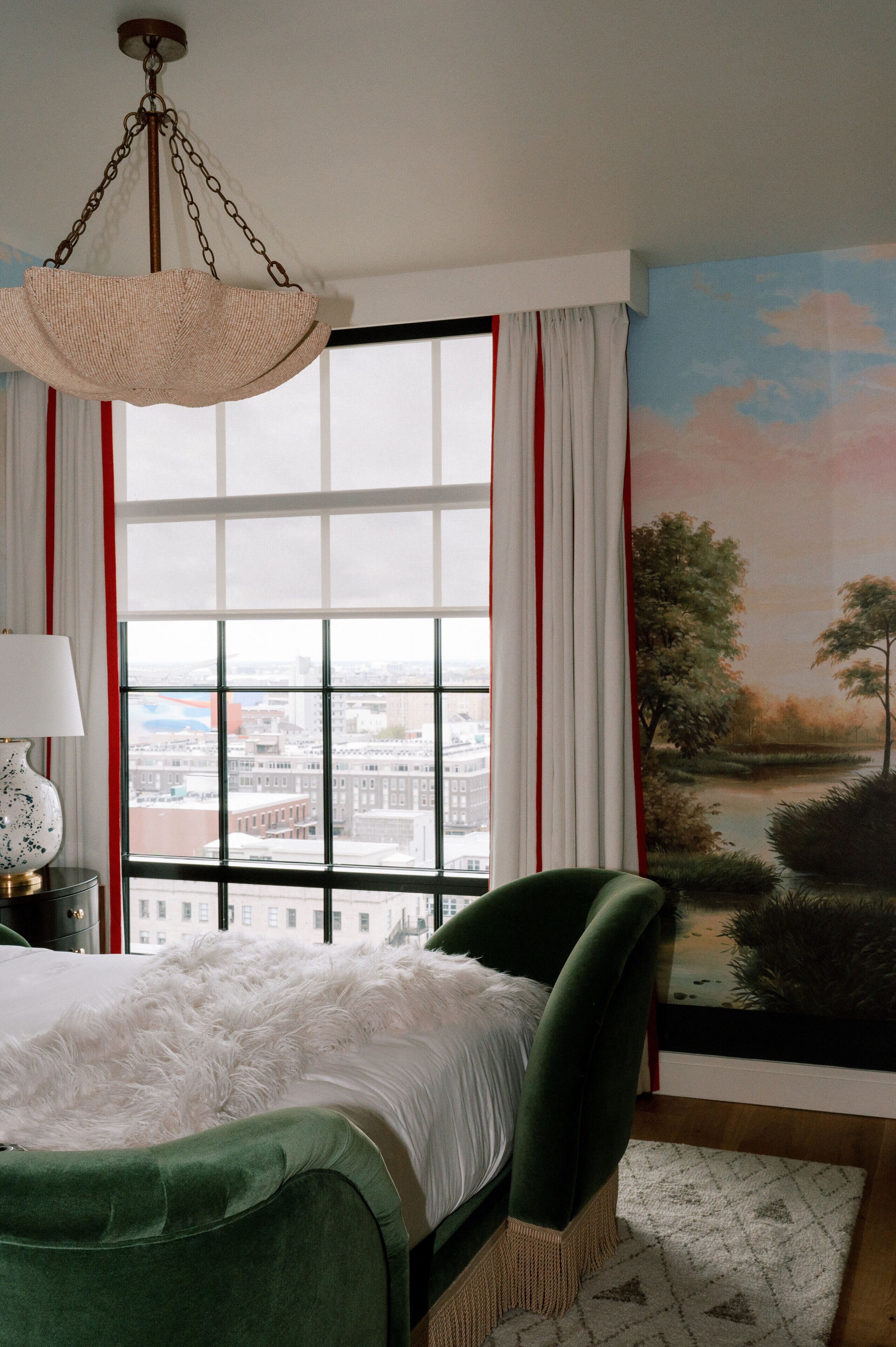 Un dormitorio con un ventanal grande, una cama con cabecera verde, un sillón verde y una lámpara colgante.