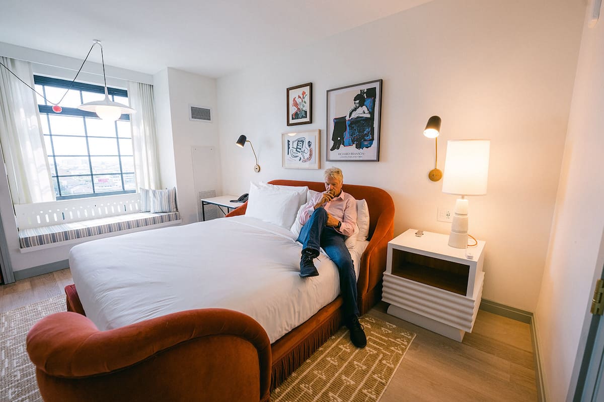 Une chambre d’hôtel avec un grand lit, une table de chevet avec une lampe, une fenêtre avec des rideaux et des œuvres d’art accrochées au mur.