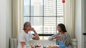 A dining area with a glass table, two people seated, and a city view through large windows.