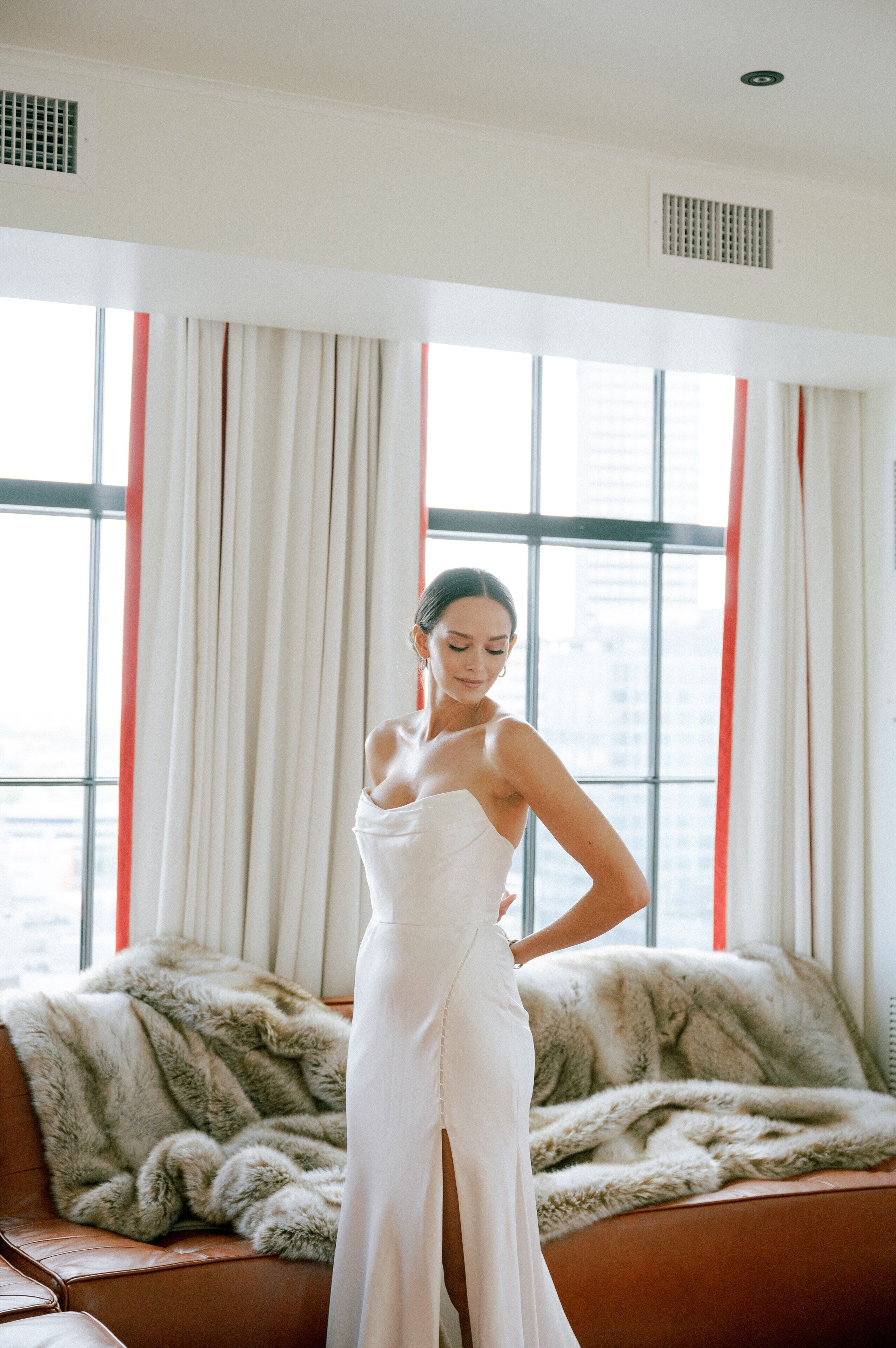 A room with a large window, a sofa with a fur throw, and a person standing in a white dress.