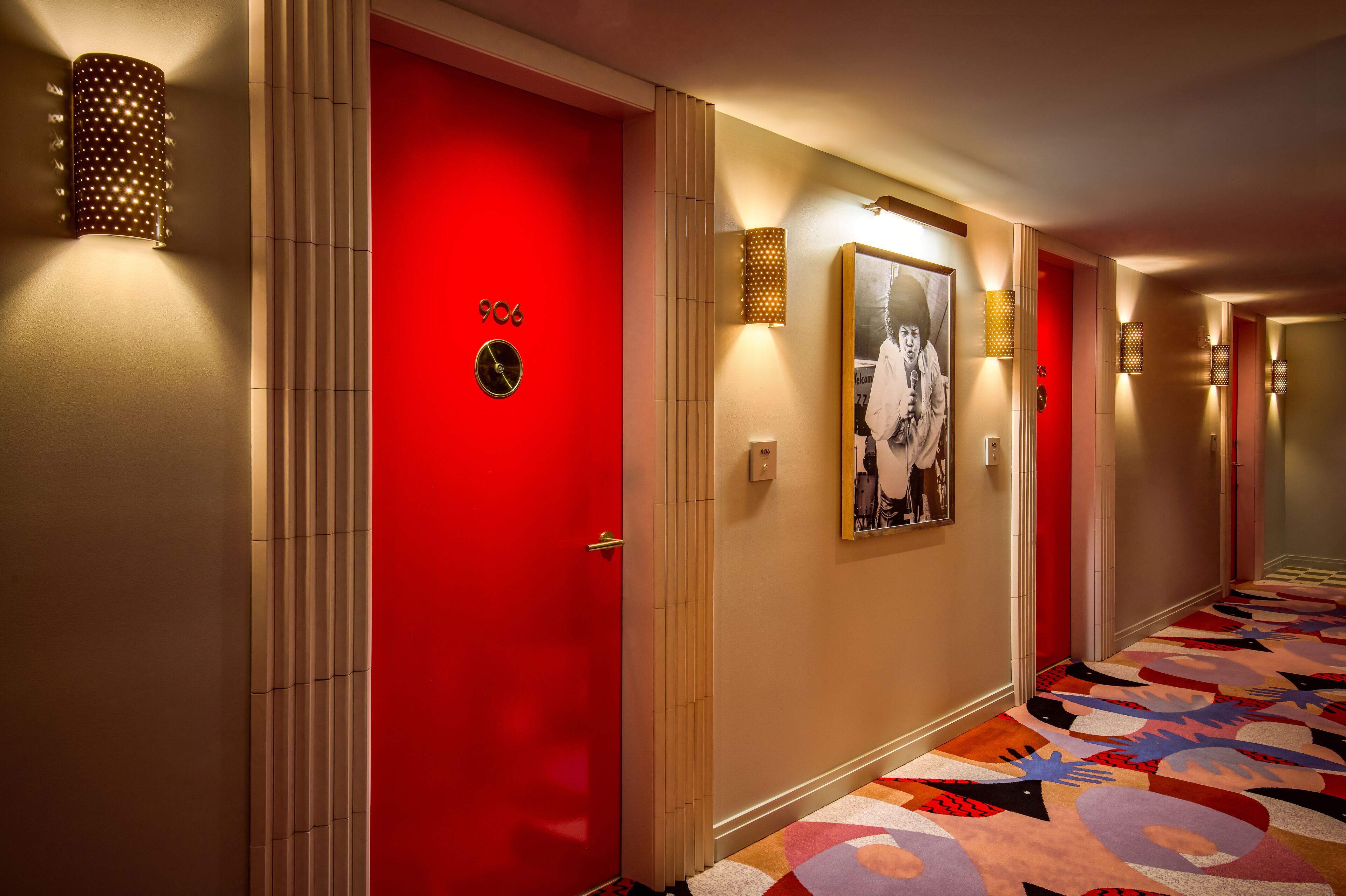 A hotel hallway with a red door, patterned wall sconces, and a colorful carpet.
