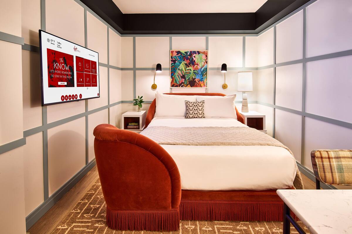 Une chambre d’hôtel moderne avec un grand lit, un fauteuil rouge rembourré, une petite table et un téléviseur fixé au mur qui diffuse une publicité rouge.