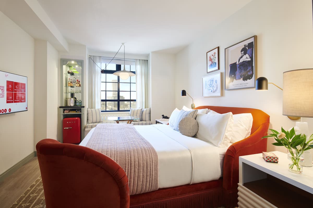 Une chambre d’hôtel avec un grand lit, un fauteuil rouge, un bureau avec une lampe, une petite table et une fenêtre donnant sur la ville.
